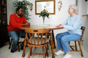 A black man in a red sweater and an older white woman dressed in blue sit and talk at a dining table.
