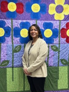 Woman with dark hair in a off white sweater posing in front of a colorful floral quilt.